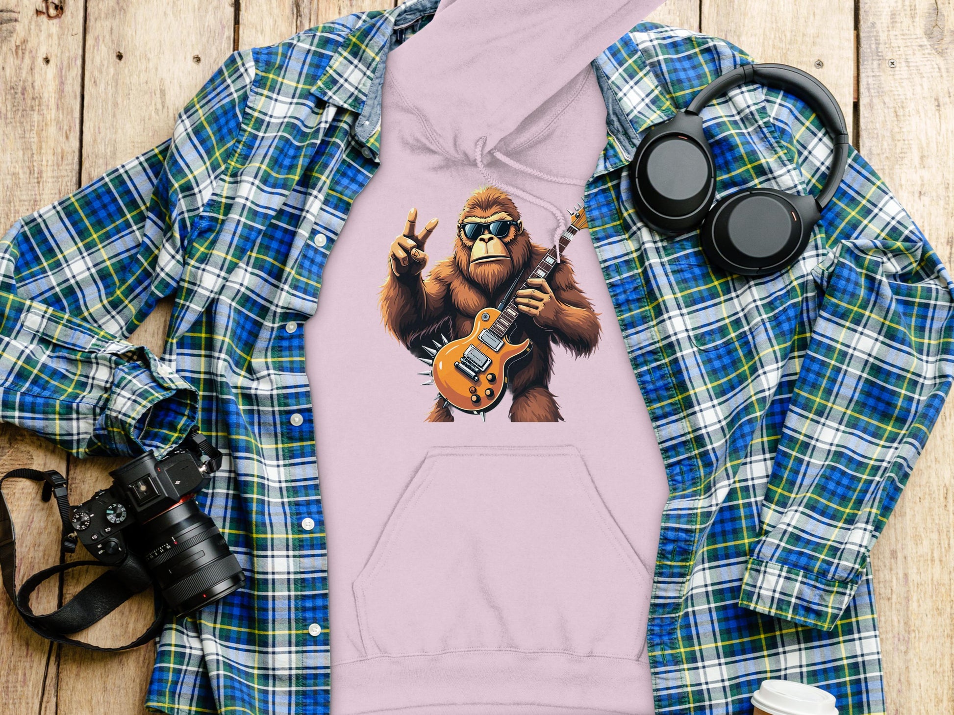 A pink hoodie from Garment Graphics, showcasing a guitar-playing gorilla in sunglasses and a green plaid shirt, sits on a wooden surface. Ideal for animal and music fans, its accompanied by a blue plaid shirt, camera, headphones, and white cup.